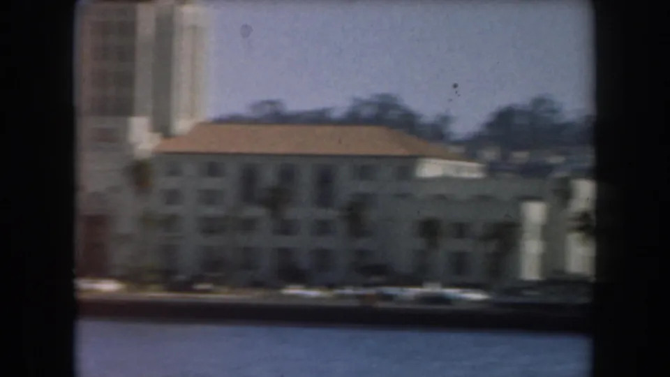 1961 California home-movie frame showing pale waterfront buildings and trees beyond blue water, viewed from a moving boat.