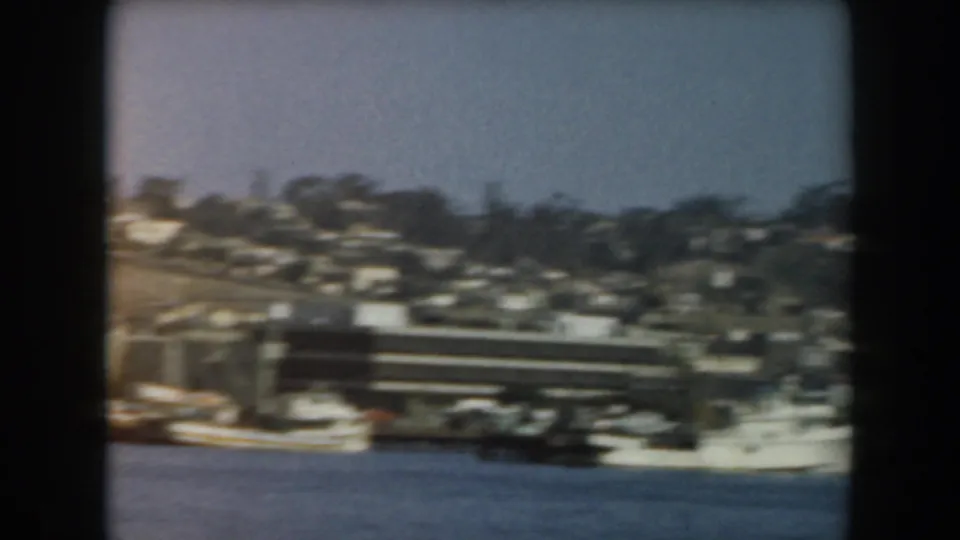 1961 California boat-view frame showing a small harbor vessel, waterfront structures, and hillside housing in soft 8mm color.