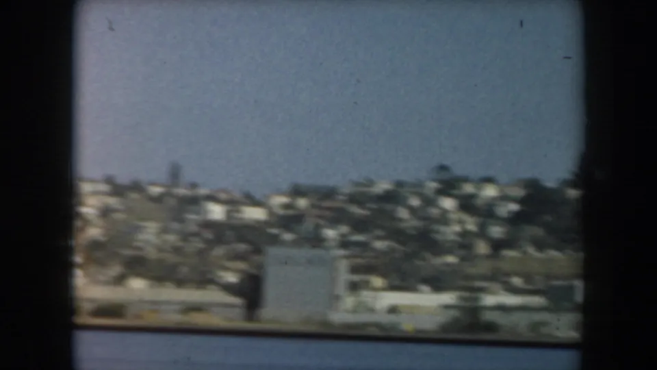 1961 California coastal frame showing open water in the foreground and a hillside neighborhood along the shore.