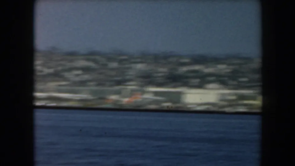 A soft 1961 California coast view shows shoreline buildings beyond blue water, partly blocked by dark framing.