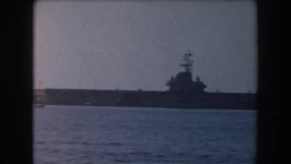 The 1961 coastal view holds on a large grey naval vessel, likely a carrier, seen from across the water.