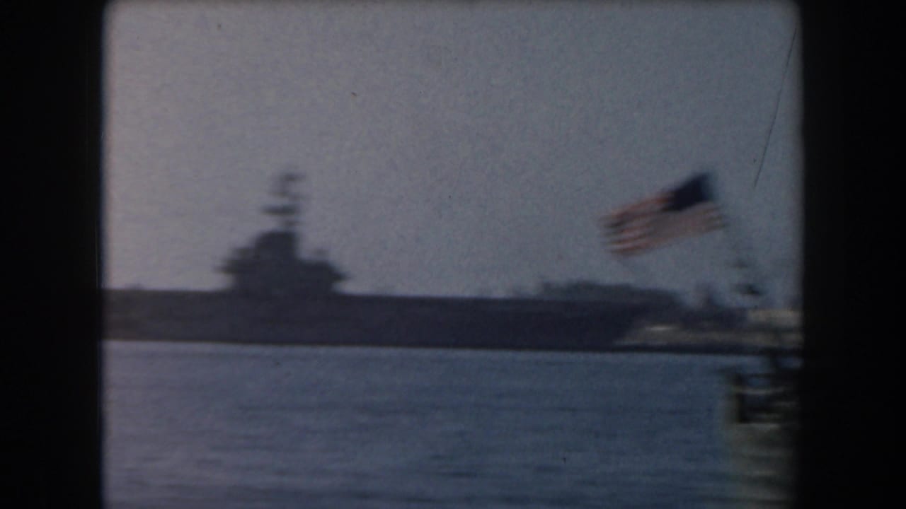 Distant aircraft carrier in harbor water with an American flag blurred in the foreground.