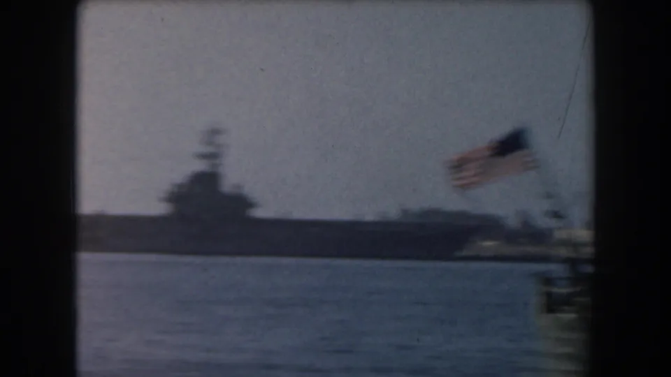 1961 California 8mm frame showing a distant aircraft carrier across harbor water with a wind-blurred American flag in front.