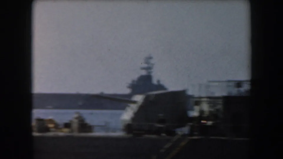 Soft 1961 California 8mm frame of a distant aircraft carrier across harbor water, partly blocked by nearby deck structures.