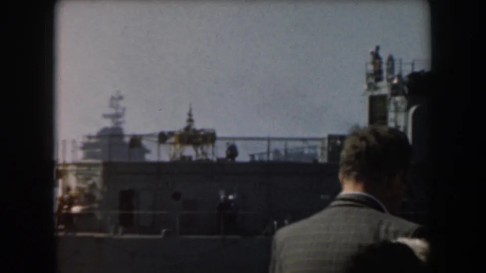 1961 California harbor frame with a suited man viewed from behind on a deck while a distant aircraft carrier sits across the water.