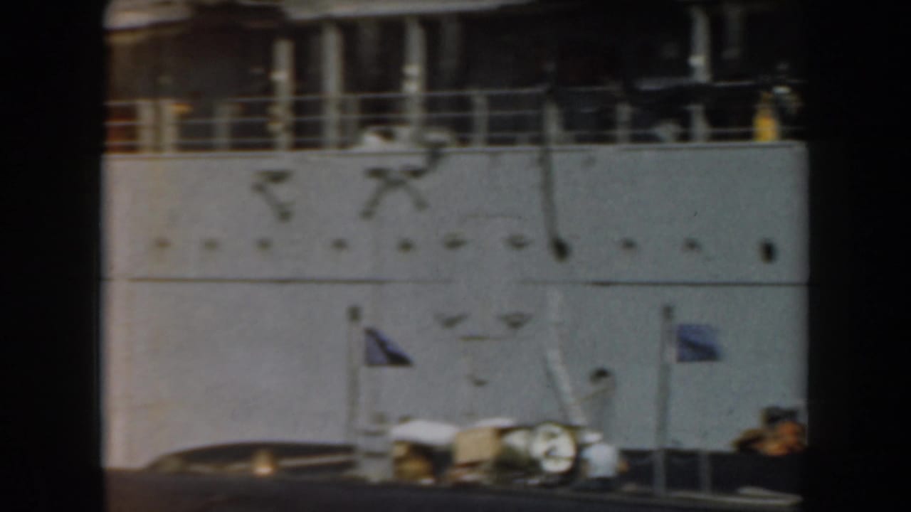 Grey naval ship side with railings, a small flag, and deck equipment in bright light.
