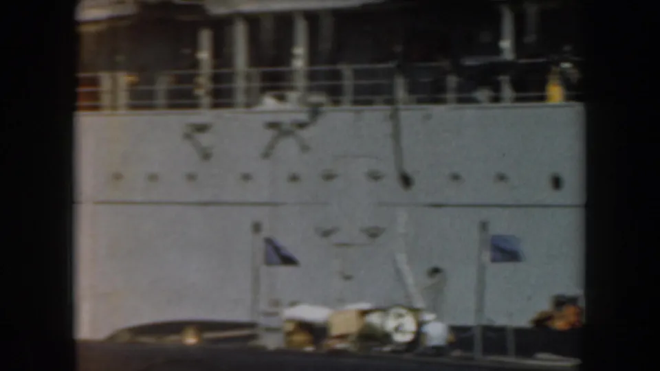 1961 California home-movie frame showing the side of a grey naval ship with railings, deck fittings, a small flag, and soft 8mm blur.
