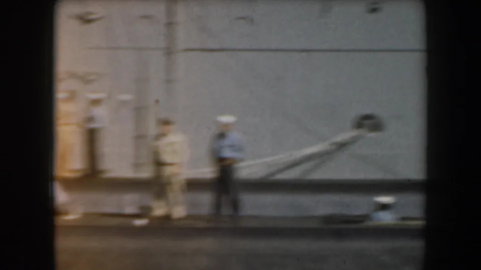 1961 California home-movie frame showing two uniformed people near a grey naval hull, with mooring lines and ship fittings visible.