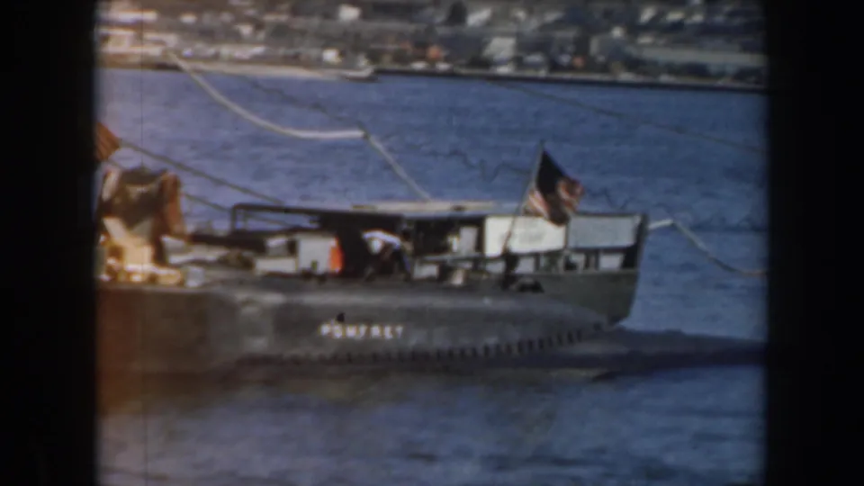 1961 California frame showing a small grey boat on blue water, with a distant shoreline and a person in a white hat on deck.