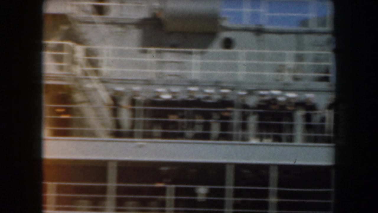 Soft 8mm frame of a naval ship exterior with deck railings and uniformed figures near the upper level.