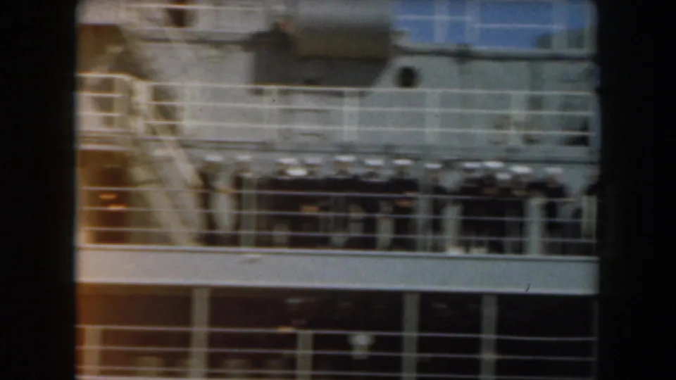 Soft 8mm frame of a naval ship exterior with deck railings and uniformed figures near the upper level.