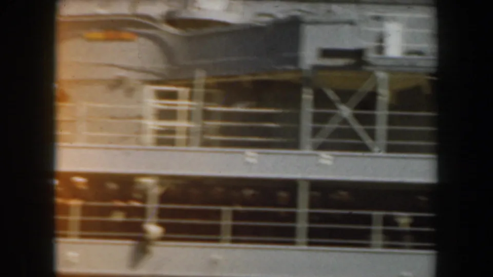 Home-movie view of a ship deck section with metal railings, ladder structure, windows, and a blurred person near the lower edge.