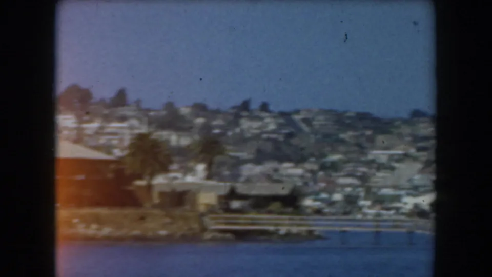 1961 California home-movie frame showing a distant hillside coastal neighborhood viewed across water from a ship.