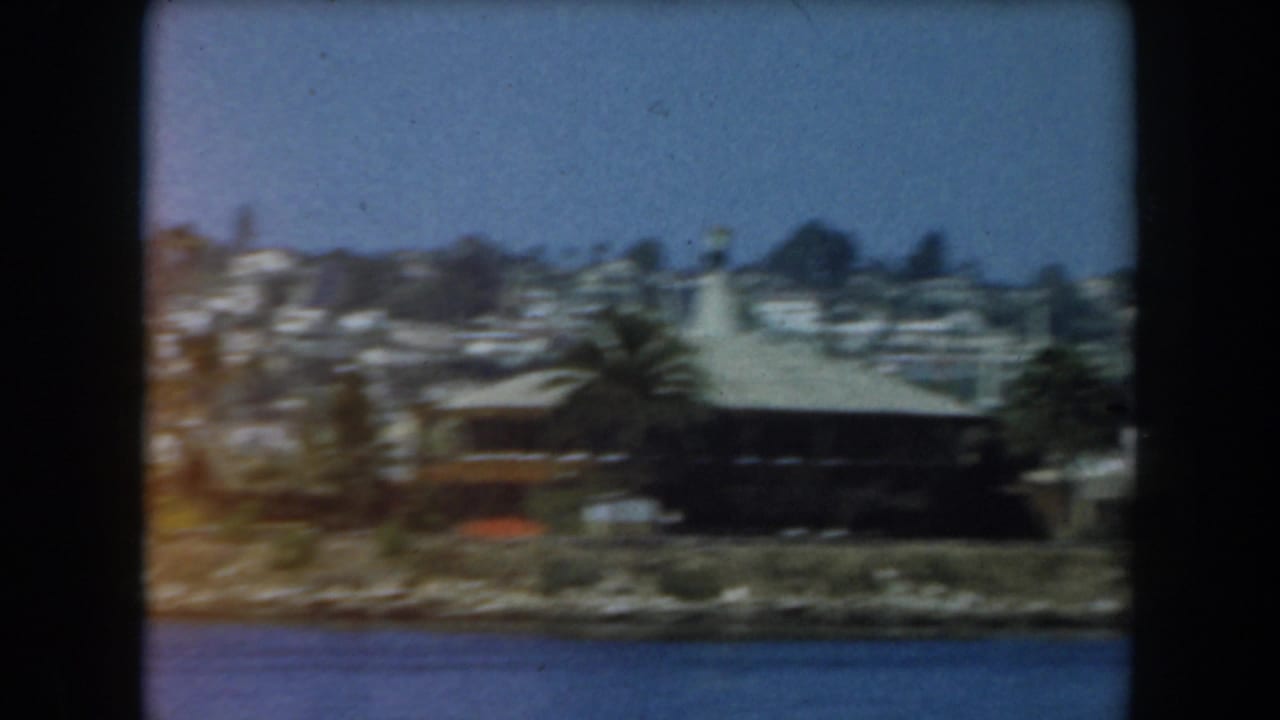 1961 California 8mm view across blue water toward a low shoreline building, palms, and homes on a wooded hillside.