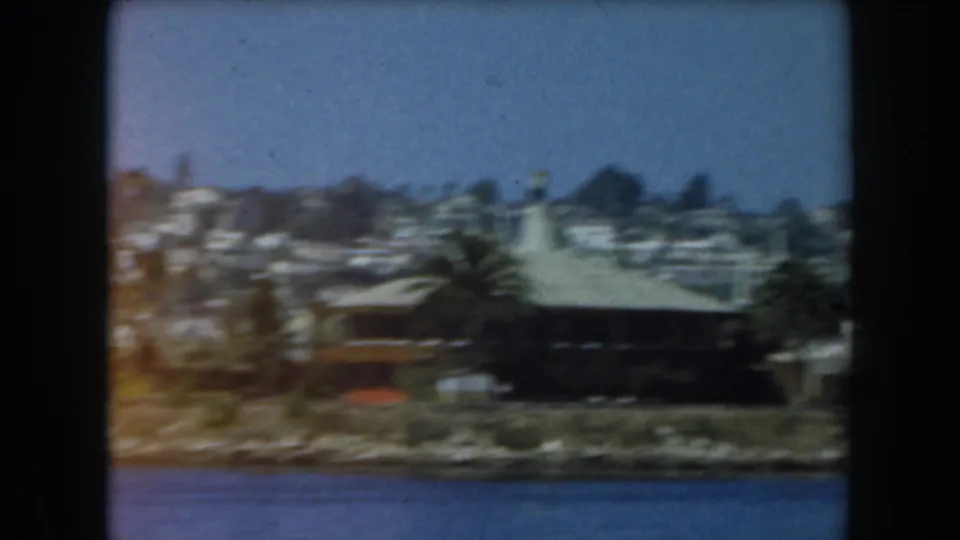 1961 California 8mm view across blue water toward a low shoreline building, palms, and homes on a wooded hillside.