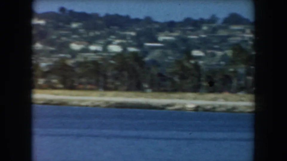 1961 California coastal view showing blue water, a pale shoreline, trees, and homes scattered across a green hillside.
