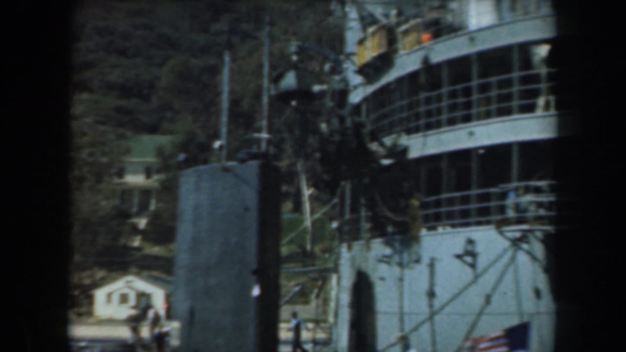 1961 California home-movie frame showing a docked white ship, dark foreground posts, hillside trees, and distant waterfront buildings.