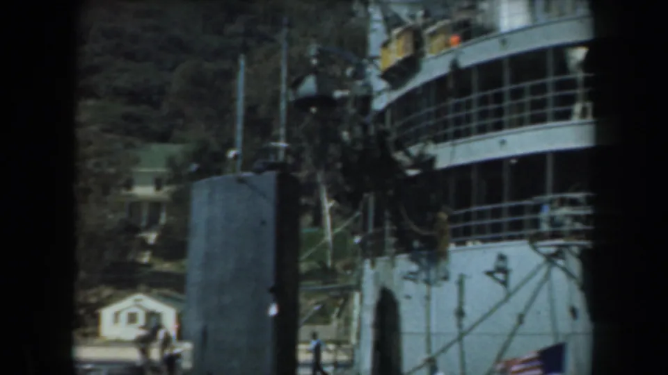 1961 California home-movie frame showing a docked white ship, dark foreground posts, hillside trees, and distant waterfront buildings.