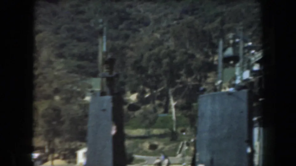 1961 California waterfront view framed by dark vertical posts, with hillside vegetation, small structures, and a ship edge visible.