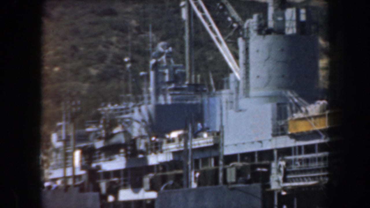 Naval vessel superstructure with cranes and deck railings near a wooded hillside.