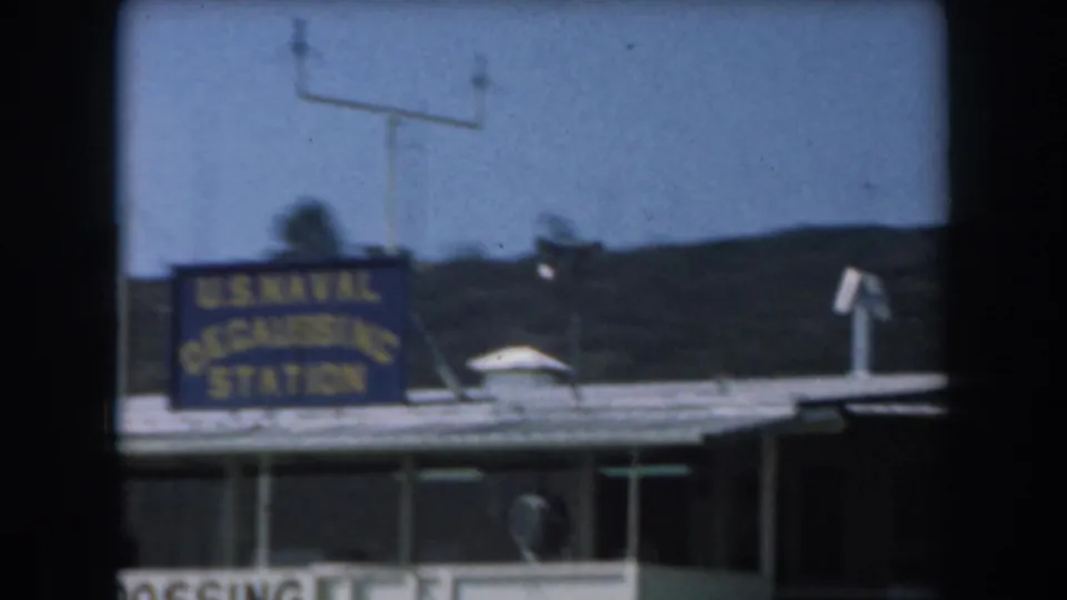 1961 California 8mm frame showing a U.S. Naval Degaussing Station sign mounted on a flat-roofed waterfront building.