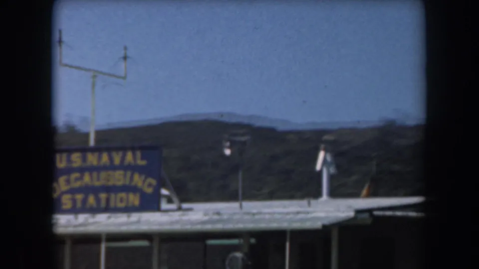1961 California 8mm view of a U.S. Naval Degaussing Station sign with a flagpole, low building, blue sky, and distant hills.