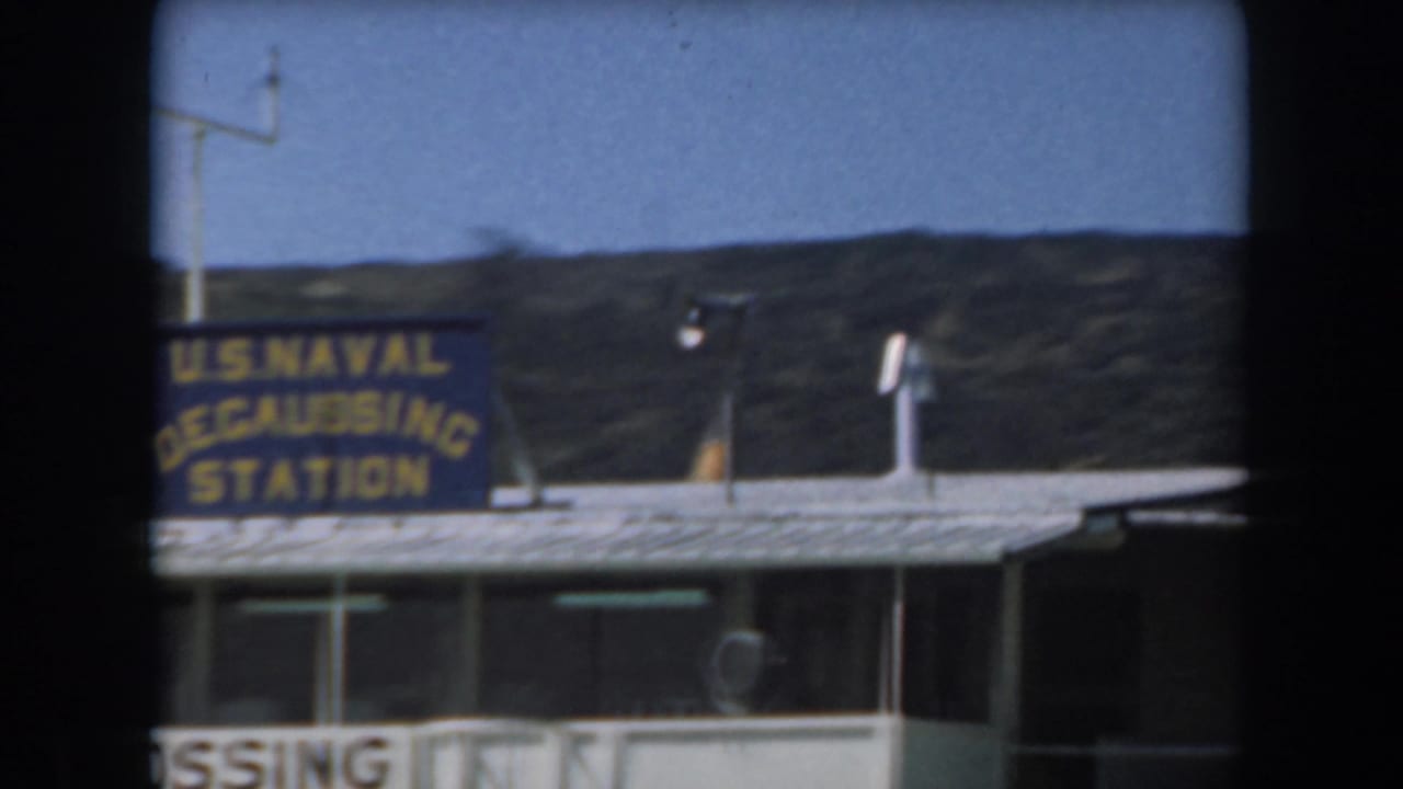 1961 California 8mm frame showing a blue U.S. Naval Degaussing Station sign on a low building against hills and clear sky.