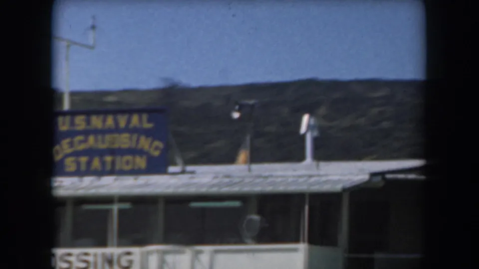 1961 California 8mm frame showing a blue U.S. Naval Degaussing Station sign on a low building against hills and clear sky.