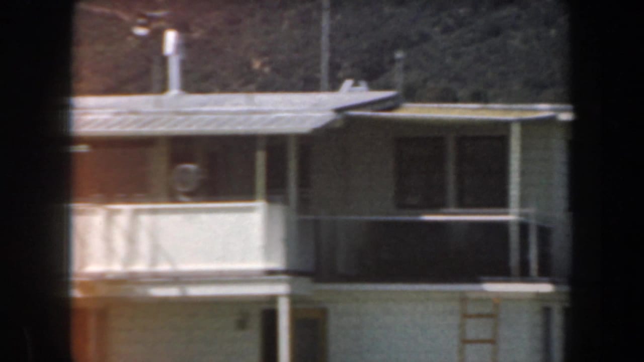 Flat-roofed building section with railing, windows, and hillside in the background.