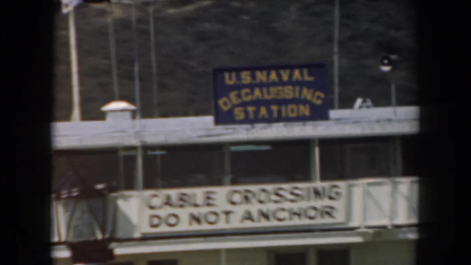 1961 California 8mm frame showing the blue naval station sign and white cable crossing warning on the facility facade.