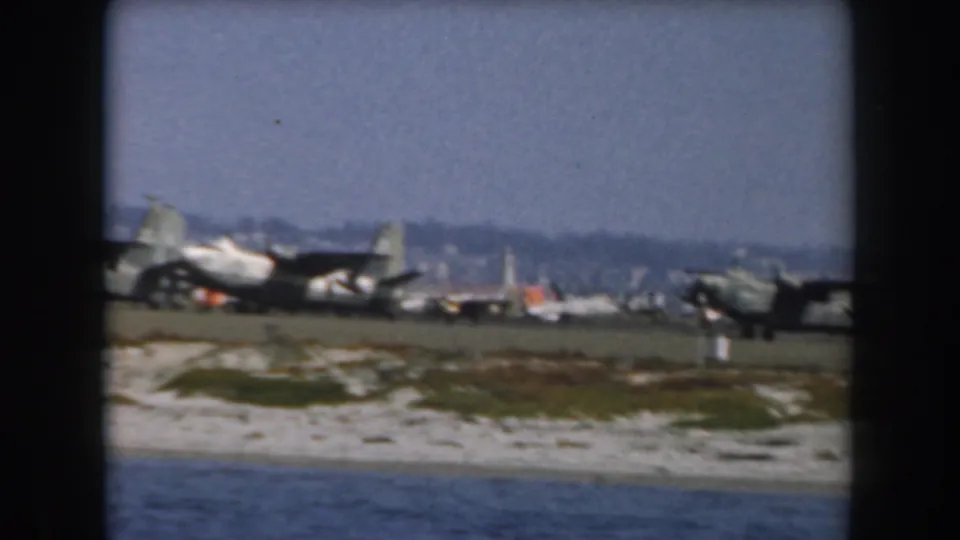 1961 California coastal view with open water in the foreground and several parked aircraft along a distant beach or airfield.