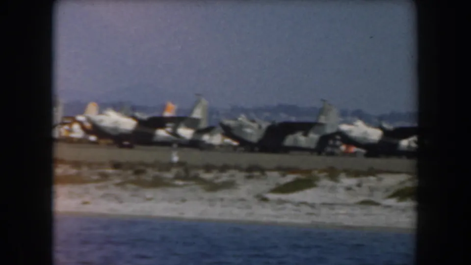 Coastal California scene showing multiple parked aircraft near a beach or shoreline, filmed across open water on 8mm.