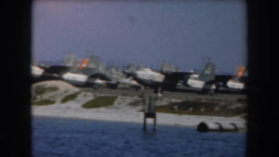 1961 home-movie frame of parked military aircraft near the sea, with a small foreground structure at the waterline.