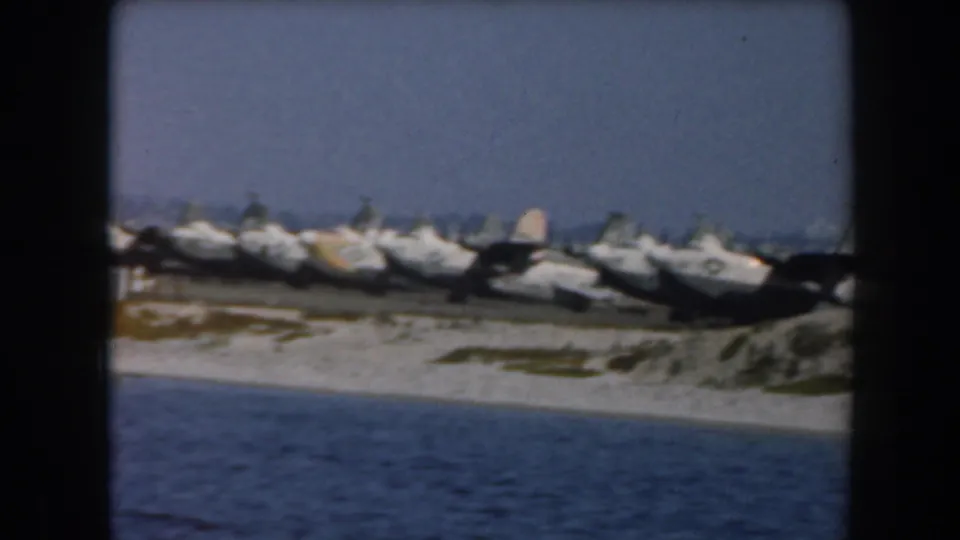 Distant 8mm view of military aircraft parked along a California shoreline, with sea water and sand in front.