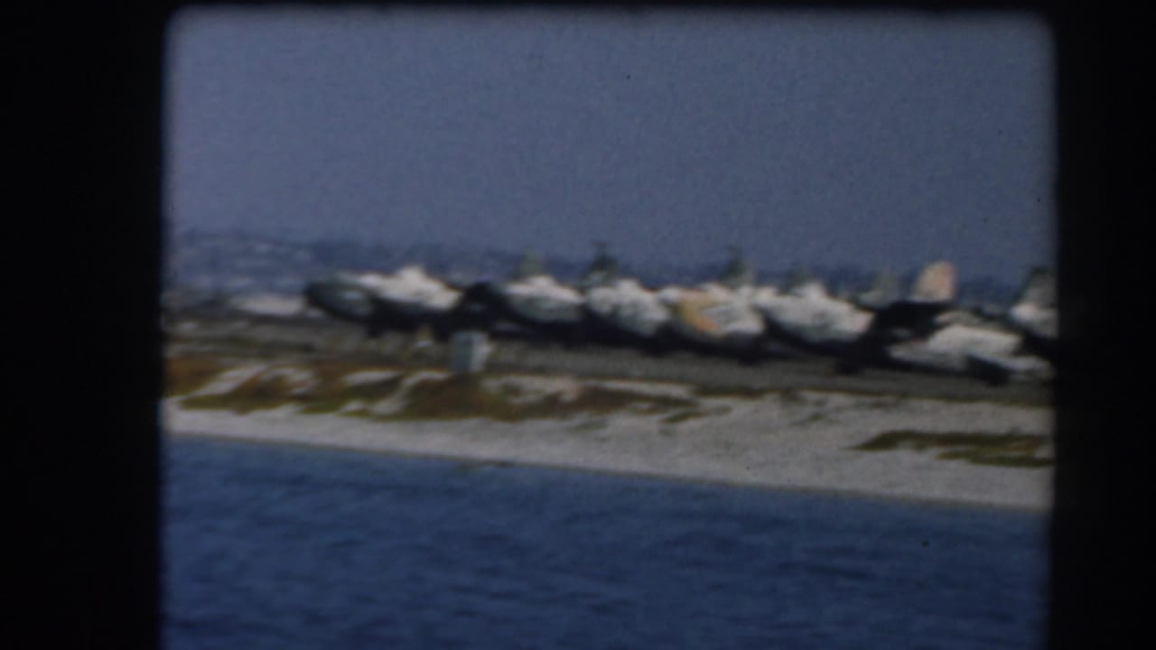 Military aircraft lined along a sandy shoreline with blue water in the foreground.