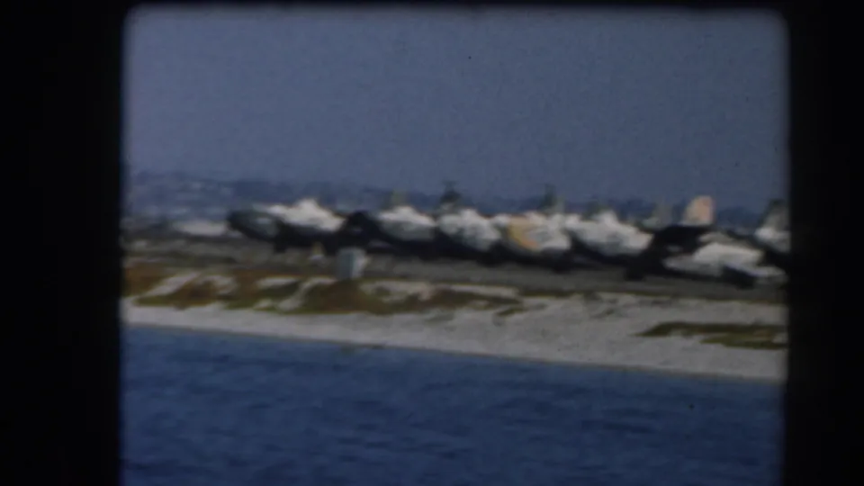 1961 California 8mm frame showing military aircraft parked near a sandy shoreline, viewed from across blue water.