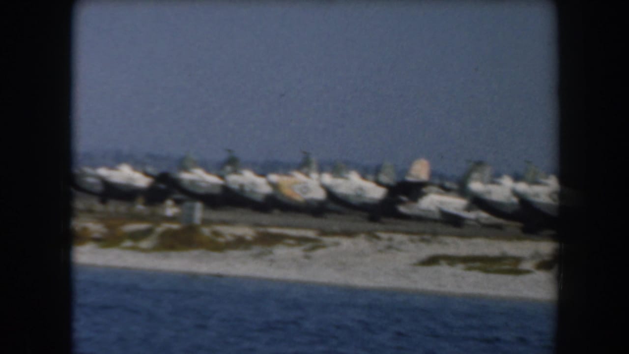 Line of military aircraft parked beside water with shoreline and sky behind them.