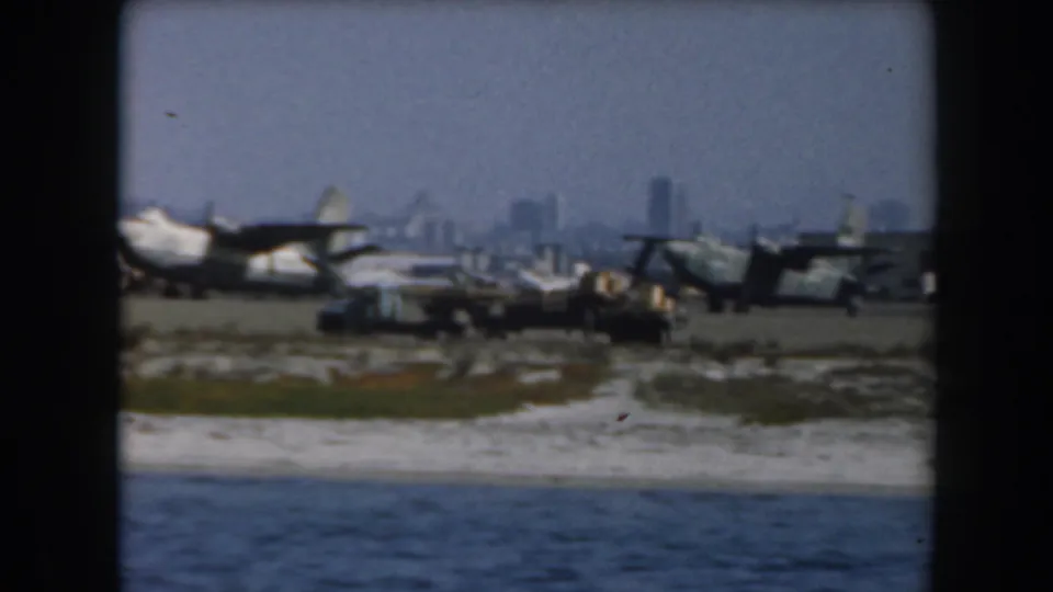 Military planes stand on runway ground near the water, with a hazy California city skyline in the distance.