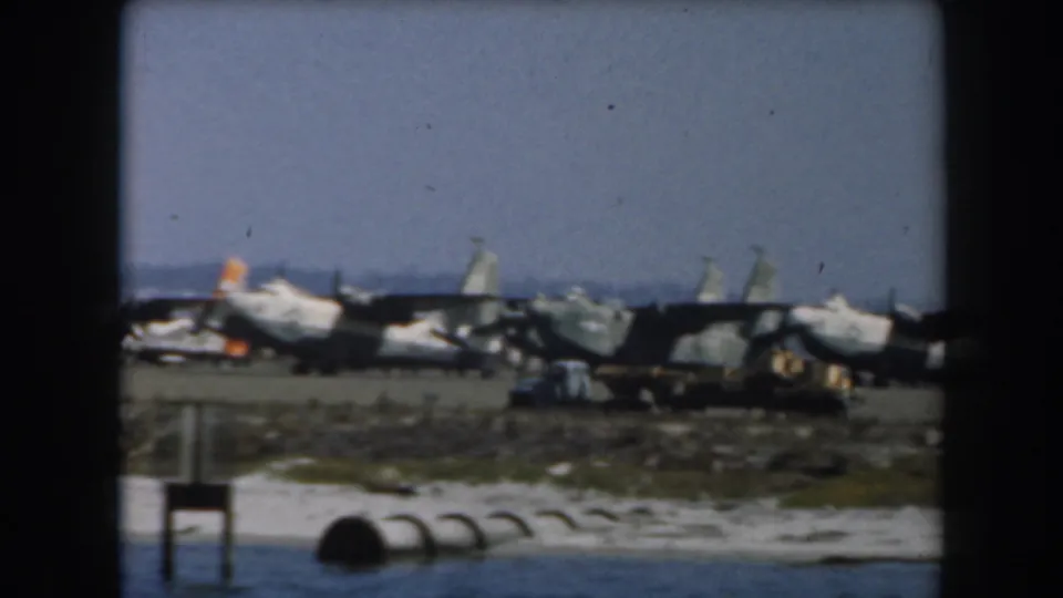 1961 California 8mm frame showing several propeller aircraft lined up on a tarmac beside water.