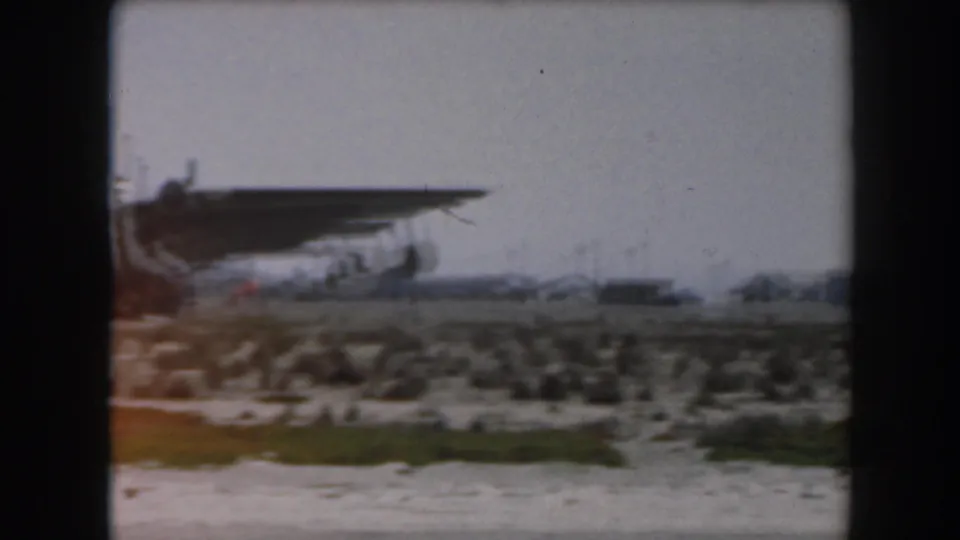 A large propeller aircraft sits across low vegetation and shoreline terrain in this 1961 California home-movie frame.