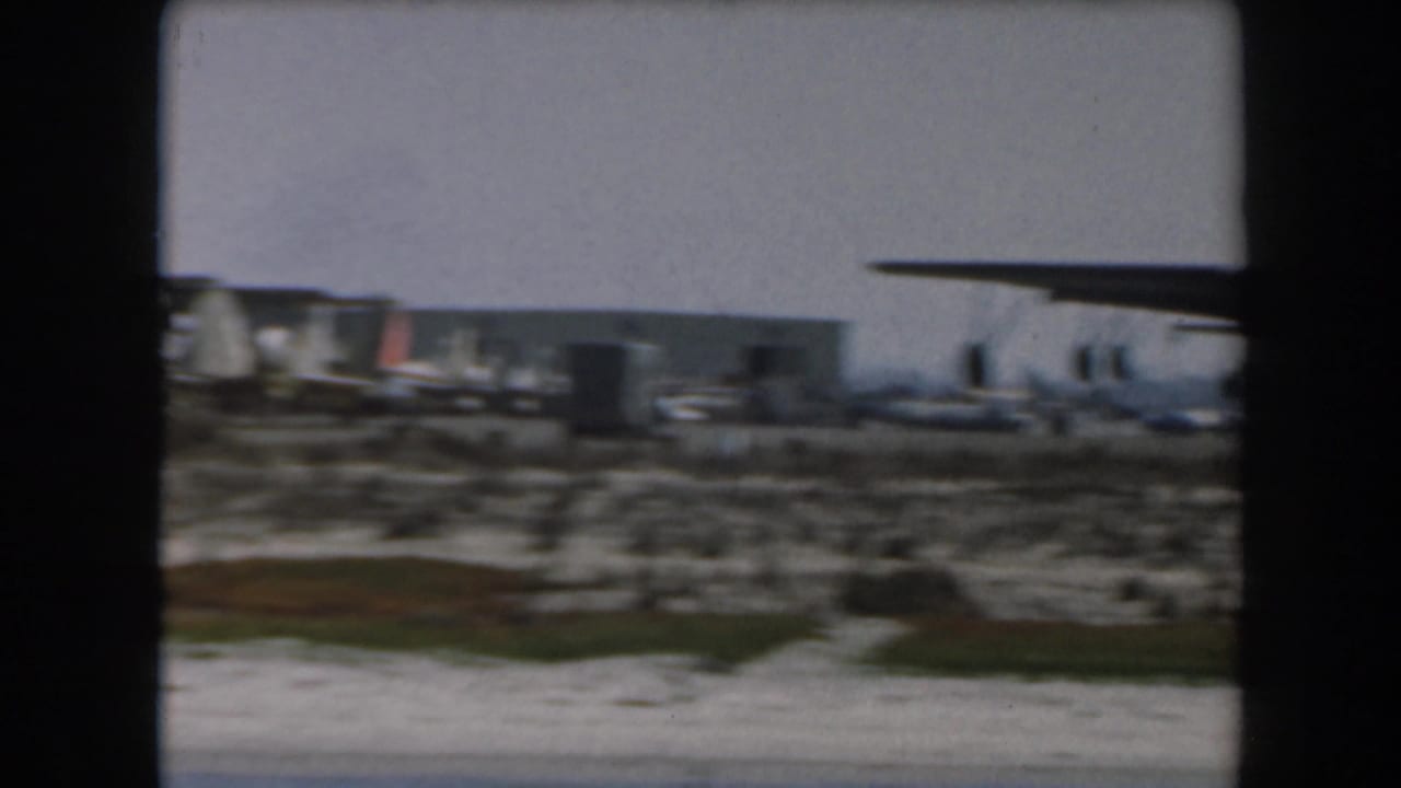 1961 California home-movie frame showing a distant large military transport aircraft on an open airfield near low structures.