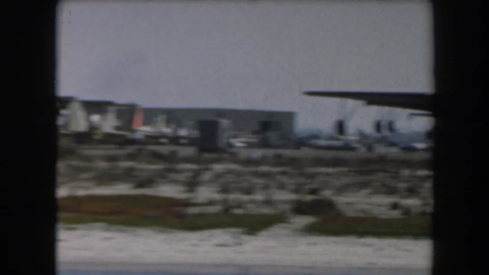 1961 California home-movie frame showing a distant large military transport aircraft on an open airfield near low structures.