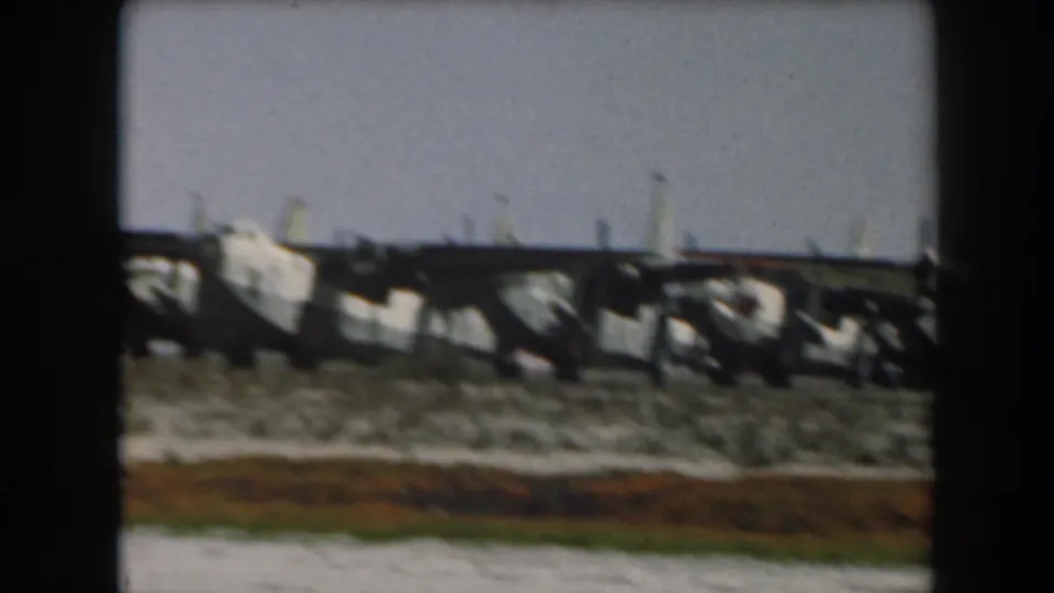 Soft 8mm frame of a distant large aircraft and low airfield structures in California, with scrub grass and open sky.
