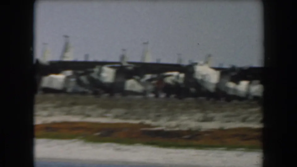 1961 California 8mm frame showing damaged rail cars on a sandy coastal track beyond a strip of water.