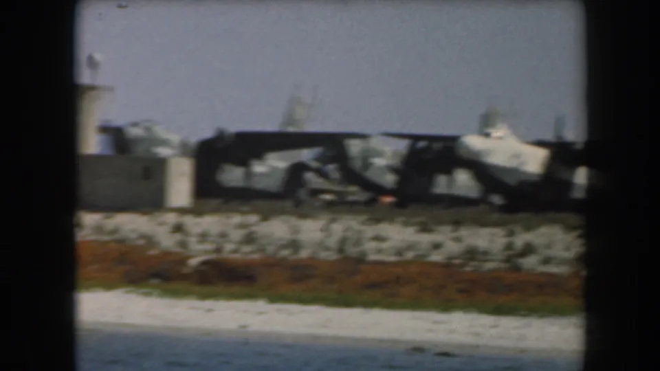 1961 California 8mm frame with derailed train cars, debris, and shoreline vegetation seen from across water.