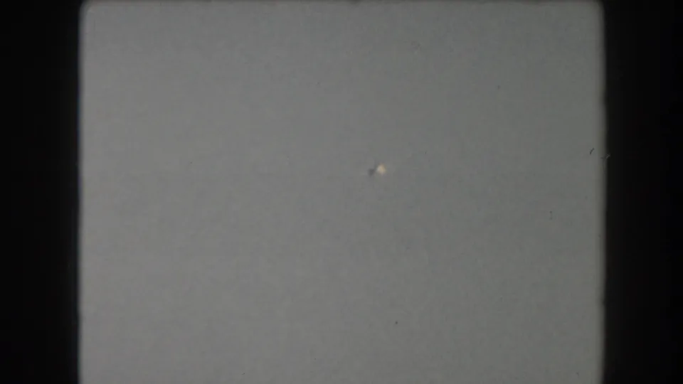 Overexposed or empty 8mm frame with a pale field, visible grain, and a small bright speck rather than clear subject detail.