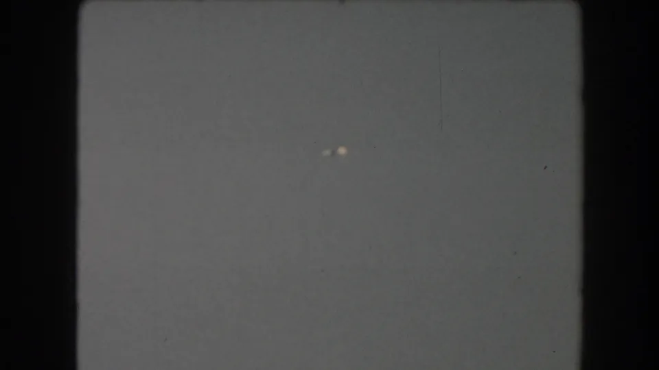 A nearly blank 8mm frame with dark film borders, faint dust, and small bright specks before the ship view begins.