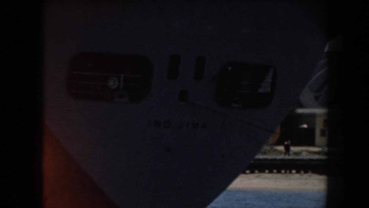 Bow of a ship with porthole openings and faint hull lettering beside a dock.