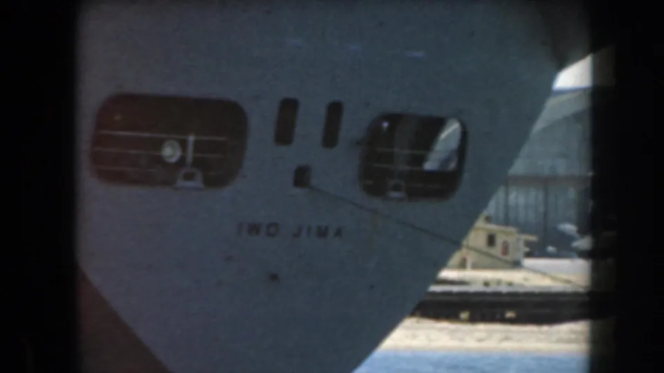 A closer 8mm frame of the vessel bow marked IWO JIMA, with hull openings, dock structures, and water visible behind it.