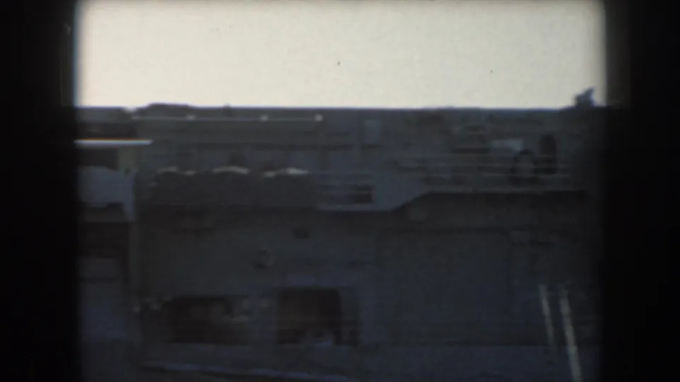 1961 8mm frame showing a utilitarian industrial passage or wall with pale panels, shadowed lower areas, and a bright opening above.